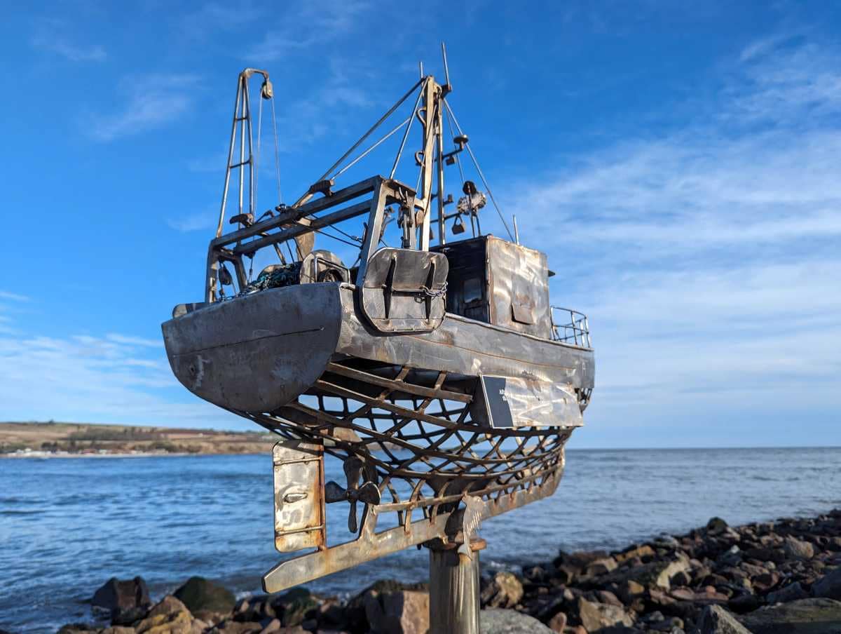 Metal boat sculpture by a rocky coast under a blue sky.